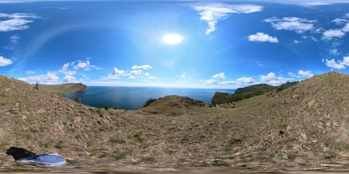 a male tourist walks on the top of a mountain on Lake Baikal, ahead is a panorama of clear blue water, a ship is sailing by, clouds in the blue sky, a view from above. 4K VR 360 degree virtual video