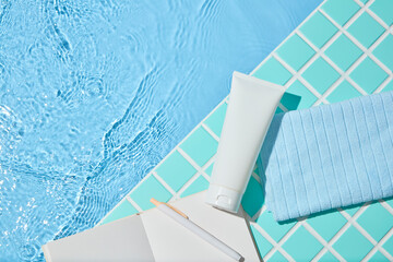 A minimal summer poolside layout with a white cosmetic tube, folded blue towel, and book. Set on...
