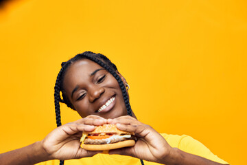 Joyful young woman of African descent holding a delicious burger against a vibrant yellow...