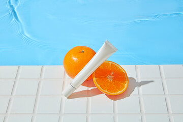 A white cosmetic tube rests beside fresh orange slices on white mosaic tiles by a clear pool....