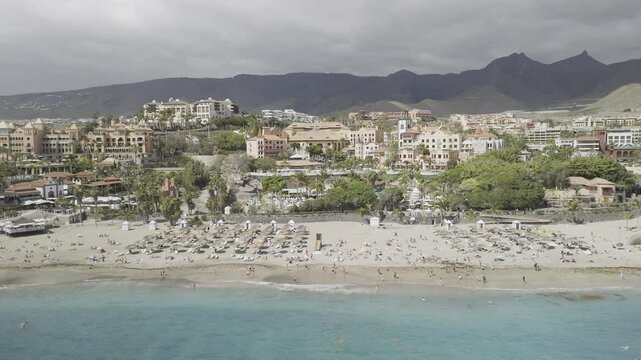 Drone flies to the left over water facing Playa del Duque beach on cloudy day in Costa Adeje, Tenerife, Canary Islands, Spain