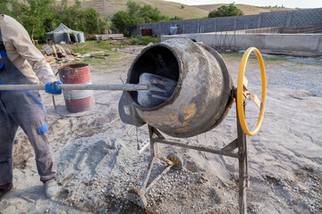 Man is using a cement mixer to mix cement. The cement mixer is yellow and has a black bucket. The man is wearing a blue apron