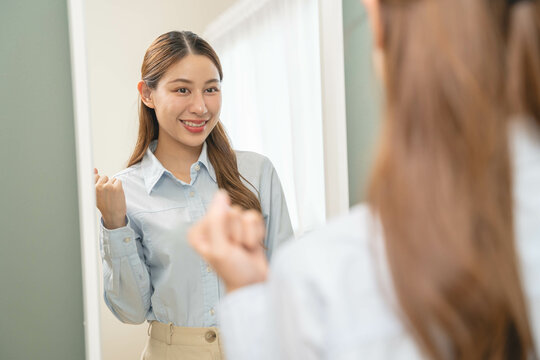 Get dress, pretty asian young woman, businesswoman standing wearing striped shirt, female getting dressed preparing before go to work looking reflection in the mirror in the morning at home.
