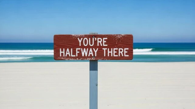 A wooden sign on a sandy beach proclaims youre halfway there under a clear blue sky, symbolizing progress and motivation