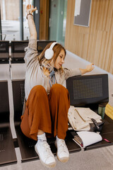 Young stylish woman in casual outfit adjusting headphones while sitting in modern public, airoport,...