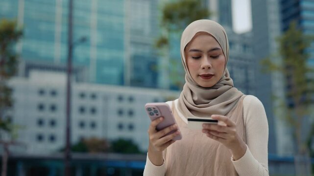 Portrait of Muslim woman wearing hijab pays online uses credit card and smartphone on city street. Malaysian lady transfers money safely via banking app in urban environment