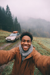 A young African man with curly hair smiles while taking a selfie outdoors. A car is parked nearby in a foggy forested area.