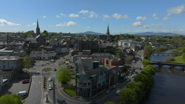Enniscorthy, County Wexford, Ireland, April 2025. Drone establishing ascend and pull away above roundabout leading to castle and tower in quaint village town on sunny day along edge of River Slaney.
