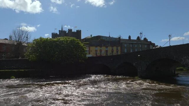 Enniscorthy, County Wexford, Ireland, April 2025. Drone tracking left over River Slaney water flowing below bridge ascending to reveal castle towers on sunny day, backlit silhouette.
