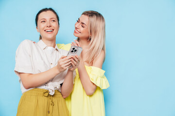 Two young beautiful smiling brunette hipster female in trendy summer clothes. Carefree women posing near blue wall. Positive models having fun. Cheerful and happy. Holding smartphone. Using apps