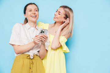 Two young beautiful smiling brunette hipster female in trendy summer clothes. Carefree women posing near blue wall. Positive models having fun. Cheerful and happy. Holding smartphone. Using apps
