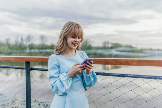 Woman in blue dress uses phone by water. Modern, outdoors.