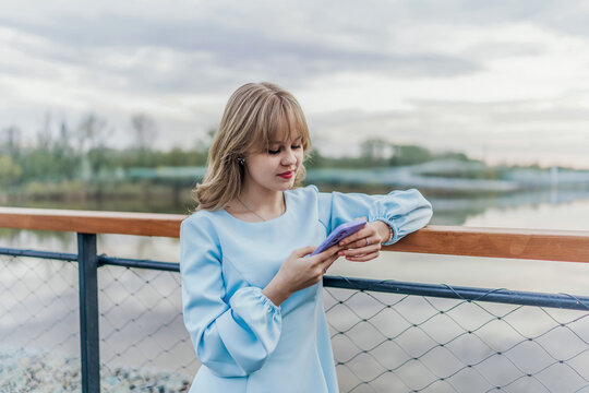 Woman in blue dress uses phone by water. Modern, outdoors.