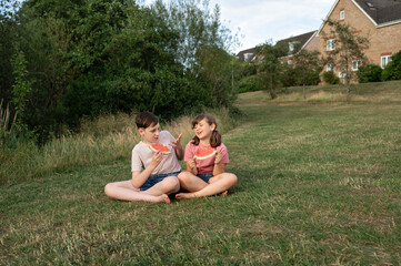 Fototapeta premium Siblings sitting in the grass, chatting and eating watermelon.