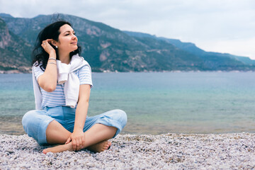 Peaceful Young Woman Sitting on Pebble Beach Enjoying Breeze and Mountain Sea View