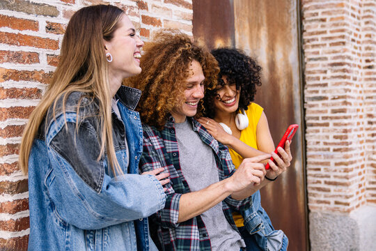 Three cheerful young friends are using a smartphone and laughing together while standing against a brick wall outdoors