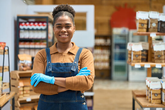 Confident saleswoman wearing gloves stands with crossed arms in grocery store