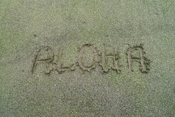 The word “ALOHA” drawn in the sand at Green Sand Beach on the Big Island of Hawaii, overlooking the turquoise bay and cliffs in the background.