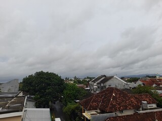 A cloudy sky over a residential area with red rooftops, green trees, and distant mountains visible...