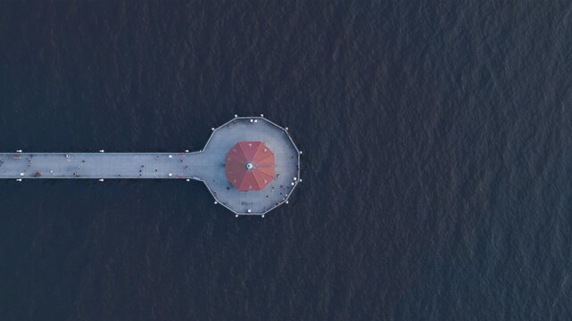 Aerial view of the Manhattan Beach Pier stretching into the dark ocean, a contrast of red and grey against the blue, Los Angeles, California, United States.