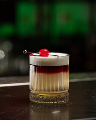 A striking layered cocktail features a rich red top over a pale yellow base, crowned with a thick...