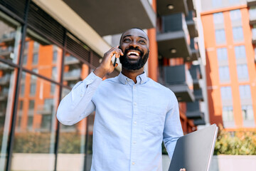 Businessman smiling and speaking on the phone while holding a laptop outside a modern building