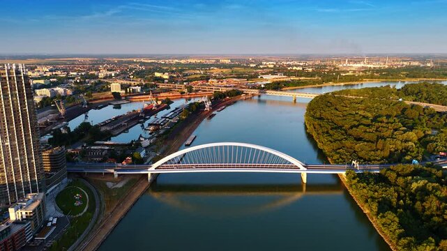 Arch of the beautiful Appolo Bridge reflects in the calm waterscape of the Danube River. Sunny view of the vast cityscape of Bratislava, Slovakia from drone.