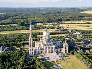 Aerial view of the Basilica of Our Lady of Lichen surrounded by lush forest, lake, and rural landscape under clear blue summer sky. Shot in Poland