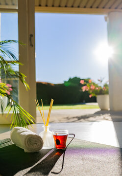 Yoga mat lying on covered porch during morning light, holding rolled towel, reed diffuser, tea mug