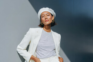Stylish woman wearing white beret and blazer posing against gray wall