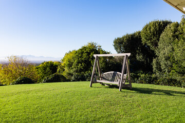 Wooden swing bench with triangle frame standing on lawn amid shrubs and mountains under clear sky
