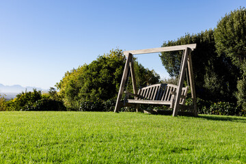 wooden swing bench is resting on green lawn under clear blue sky, shrub and tree border