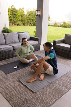Couple in sportswear stretching on yoga mats on patio overlooking lawn and reaching for smartphone