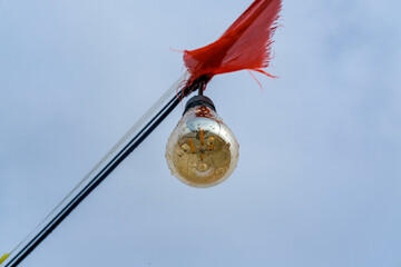 Outdoor lightbulb hanging from a wire beneath a tattered red flag, captured against a pale sky in a...