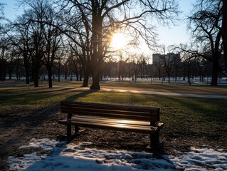A wooden bench in a park during winter with bare trees and sunlight shining through, casting long shadows on the ground with patches of snow.