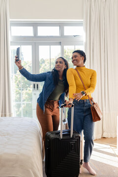 Diverse female friends posing for selfie in bright bedroom near black suitcase and smartphone