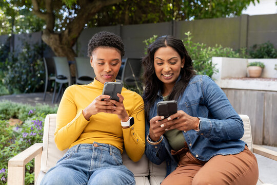 Diverse female friends scrolling on smartphones and checking smartwatches seated on bench in garden