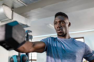 African American man wearing blue t-shirt lifting adjustable dumbbell at gym near kettlebell rack