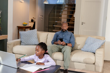 African American father and daughter using smartphone and studying at home with notebook and pencil