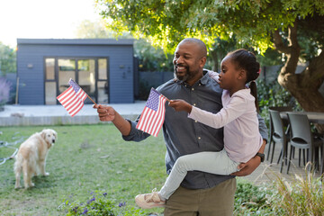 African American father and daughter standing in backyard waving American flags by golden retriever