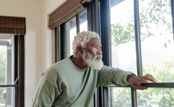 Senior African American man in sweatshirt leaning on window sill with blinds at home gazing outside
