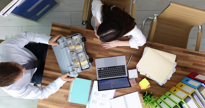 Man opens briefcase filled with money while surprised woman gesturing with hands in shock. Bribe at wooden desk with business documents in office