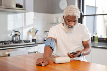 African American man checking blood pressure using monitor and phone in kitchen, copy space