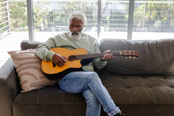 Senior African American man strumming acoustic guitar sitting on grey sofa near sliding doors