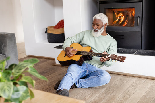 Senior African American man wearing green sweatshirt strumming acoustic guitar at home by fireplace