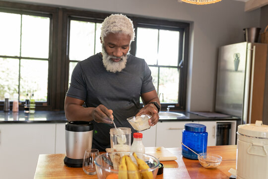 African American senior man spooning mixture from bowl into blender jar at kitchen island - Powered by Adobe