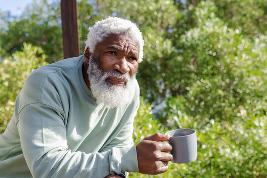 Senior African American man leaning on deck railing holding gray mug, looking ahead in garden