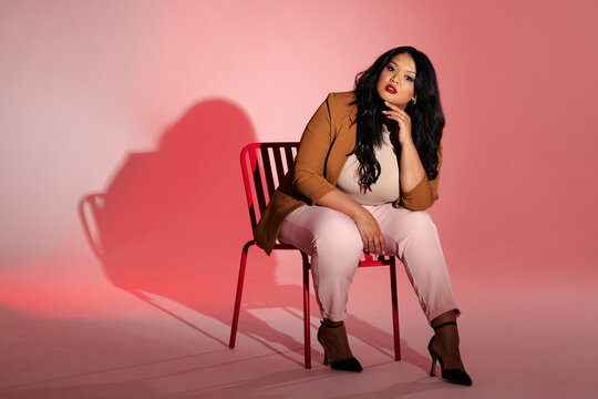 Woman sitting on red metal chair in studio resting chin on hand while wearing camel-brown blazer