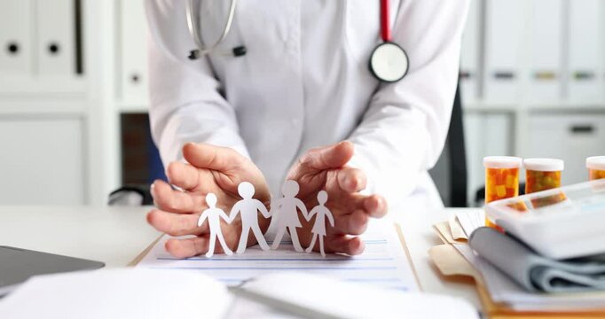 Doctor holds cupped hands over paper cut-out chain of family symbolizing protection and care. Medical office emphasizes family health and insurance