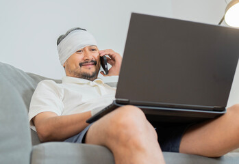 Bearded man with head injury sitting on sofa, uses laptop and phone at home for working online.